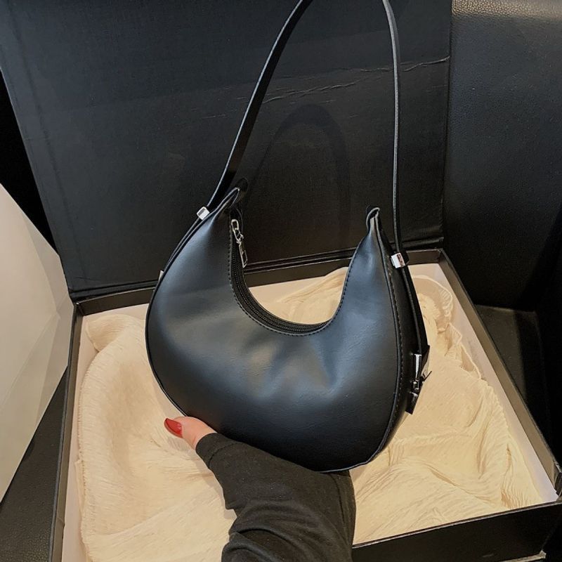 Black crescent shoulder bag held in hand, showing the zipped opening, smooth surface and silver-tone strap buckle